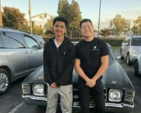 Two kids posing for a photo, smiling and leaning up against a car