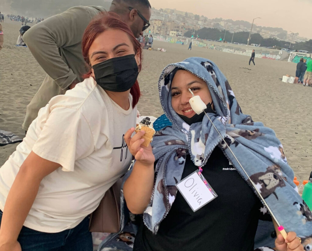 Two teens posing and smiling together. One wearing a mask and one covered in a blanket