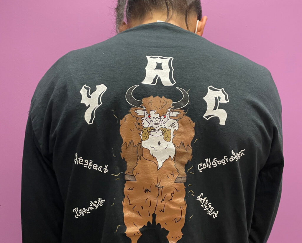 Backside view of a person wearing a shirt that says YAC on it with an illustration of a yak type creature